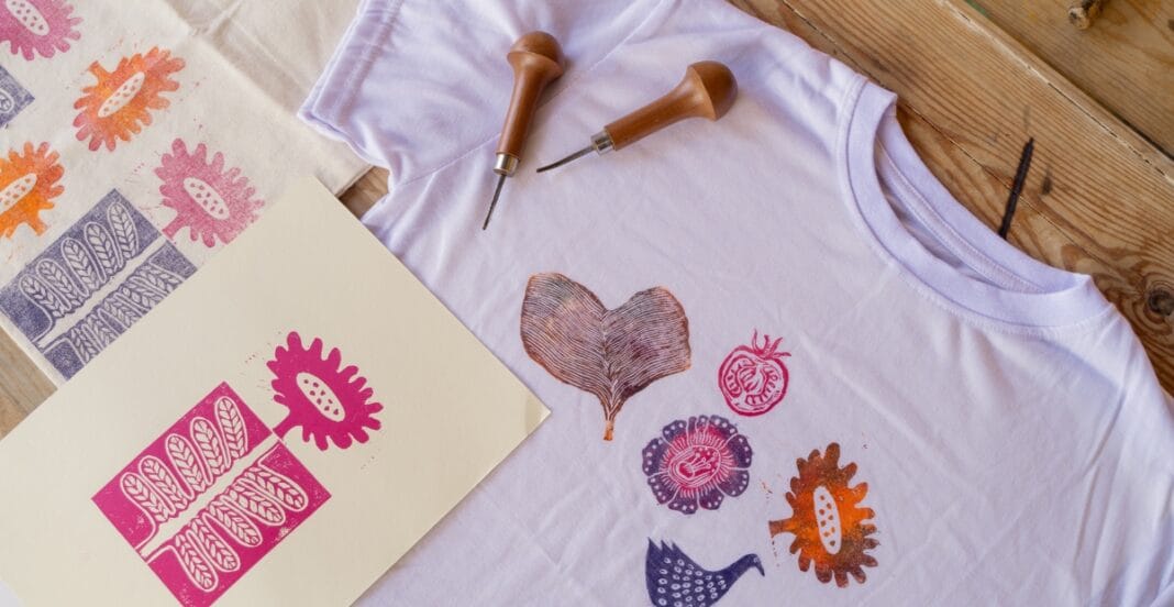 A white T-shirt spread on a wooden surface with several colorful, flower-shaped stamps printed on it. Nearby are stamping tools (handles and metal tips) and a cream-colored sheet with bold pink stamp designs. The scene suggests a DIY stamping or textile art project.