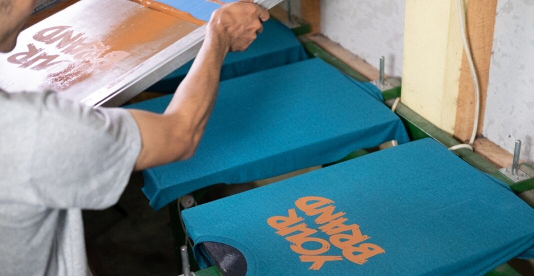 A close-up of a screen printing setup where a person presses a squeegee over a blue t-shirt on a printing rack, with orange text partially visible on the shirt.