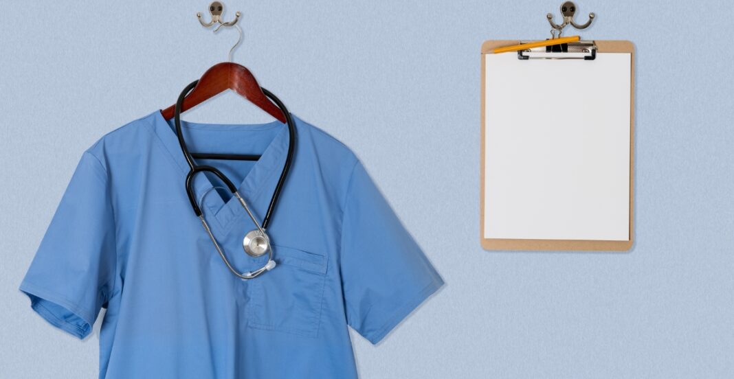 A blue medical scrub top hanging on a hanger, with a stethoscope draped around its neck, next to a blank notepad on a wall hook against a light blue wall.
