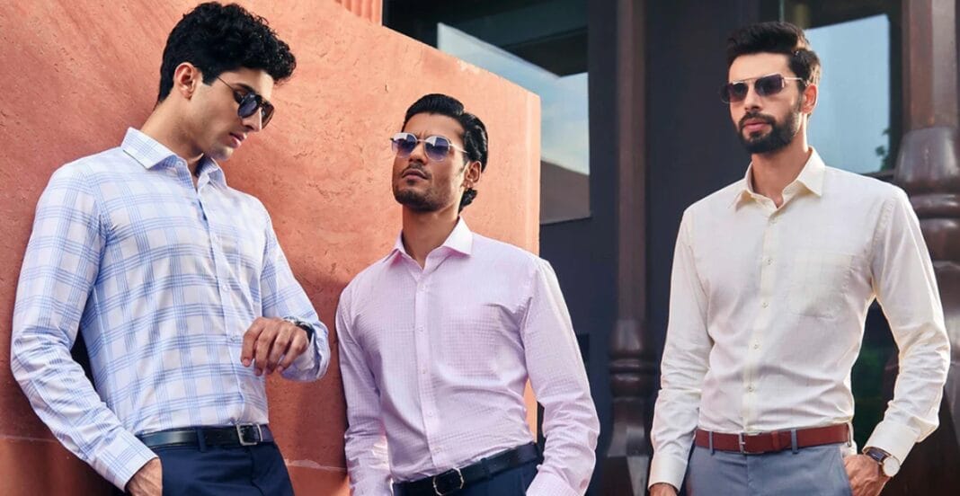 Three stylish men wearing sunglasses and formal shirts standing outdoors. Two are wearing light-colored shirts (one in white with a check pattern and the other in plain white), while the third man is in a pastel pink shirt. They are positioned near a reddish wall with modern building elements in the background.