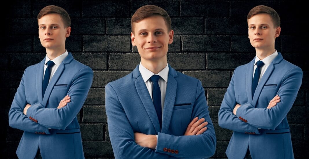 A trio of young men in matching light blue suits and dark blue ties stand against a dark brick wall, each with arms crossed. The man in the center smiles confidently, while the two on the sides look straight ahead with neutral expressions.