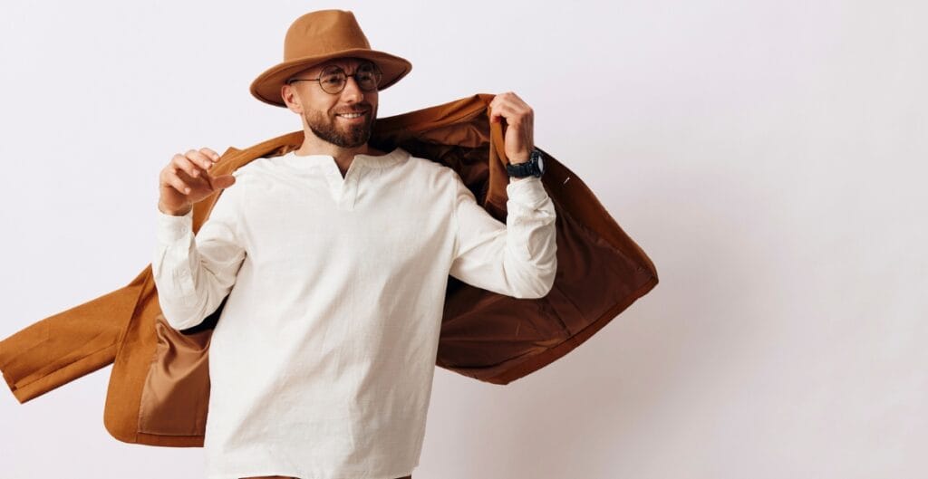 A smiling man wearing a light-colored long-sleeve shirt, a brown hat, and glasses, playfully holding open a brown jacket as if about to put it on.