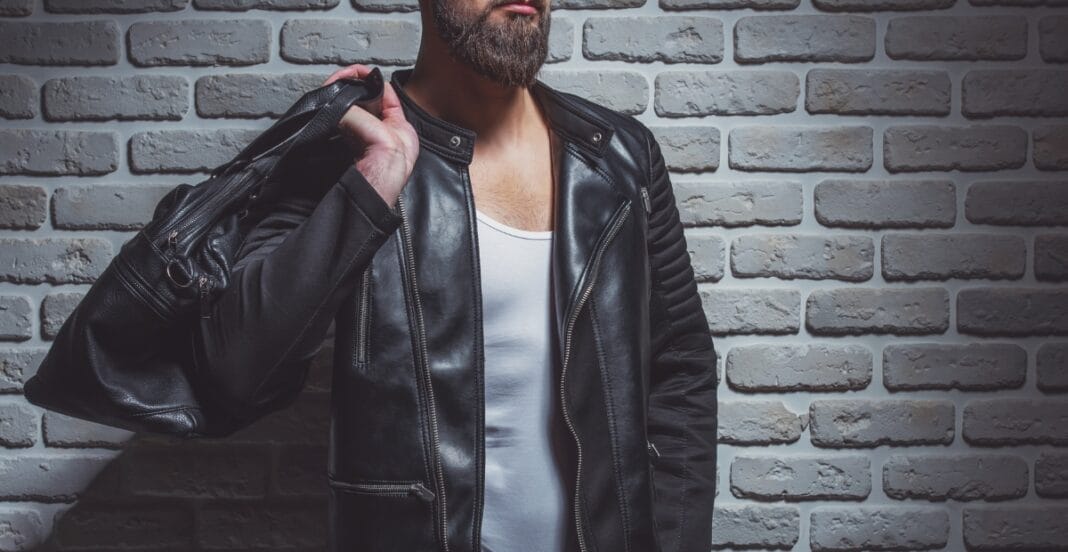 A man with a beard wearing a black leather jacket and a white shirt, carrying a black shoulder bag, standing against a grey brick wall.