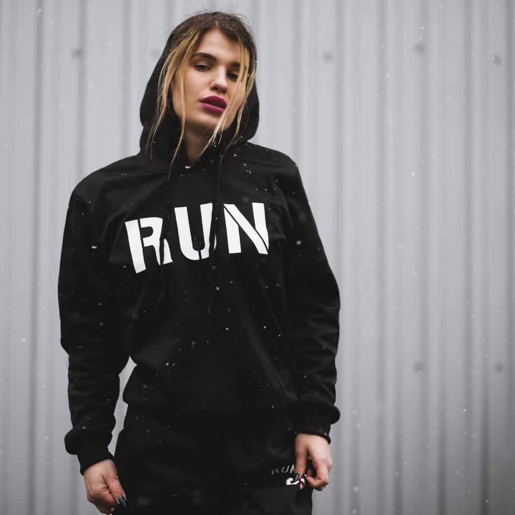 Young woman wearing a black hoodie with "RUN" written in bold white letters across the chest, standing outdoors in the rain against a metallic background. She has blonde hair with darker roots and is looking slightly down with a neutral expression.