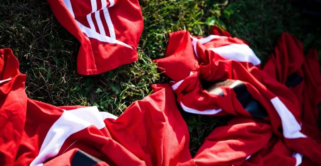 Red sports uniforms with white stripes and accents lying on green grass under sunlight.