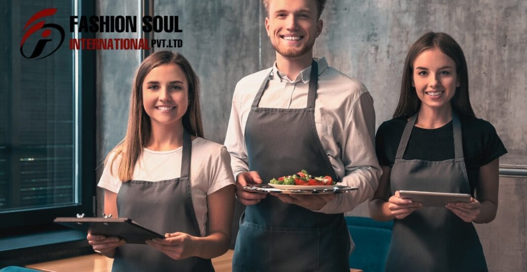 Three smiling restaurant staff members wearing aprons stand side by side indoors; the woman on the left holds a clipboard, the man in the middle carries a plate of food, and the woman on the right holds a tablet. A logo reading “FASHION SOUL INTERNATIONAL PVT. LTD” appears in the upper left corner.