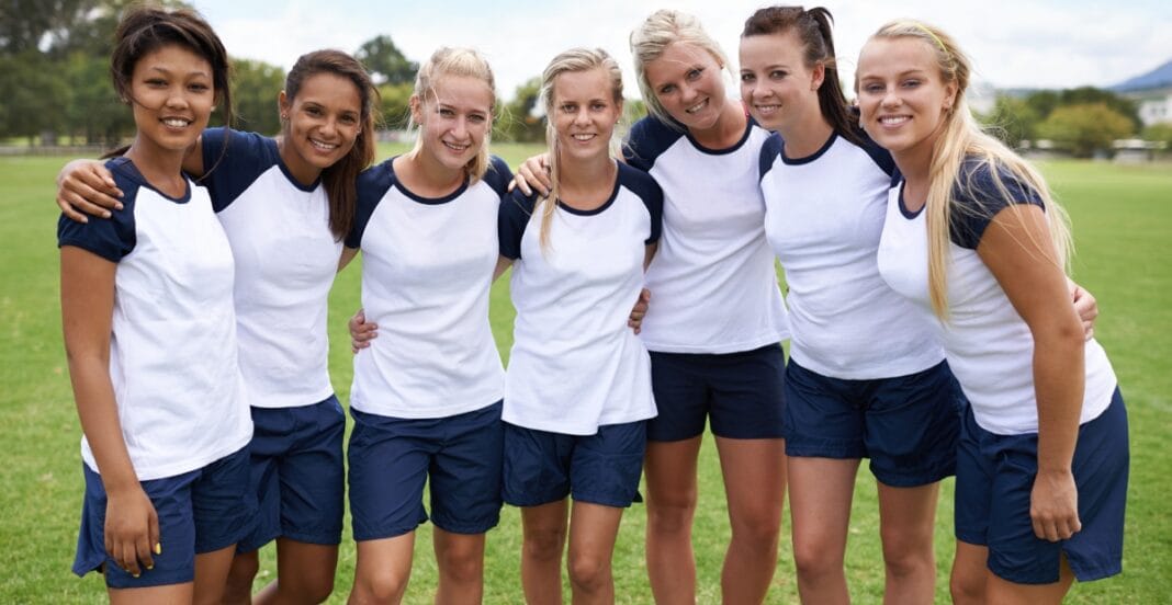 A group of seven young women standing closely together outdoors on a grassy field, smiling at the camera. They are wearing matching white and navy blue athletic shirts and shorts, likely part of a sports team.