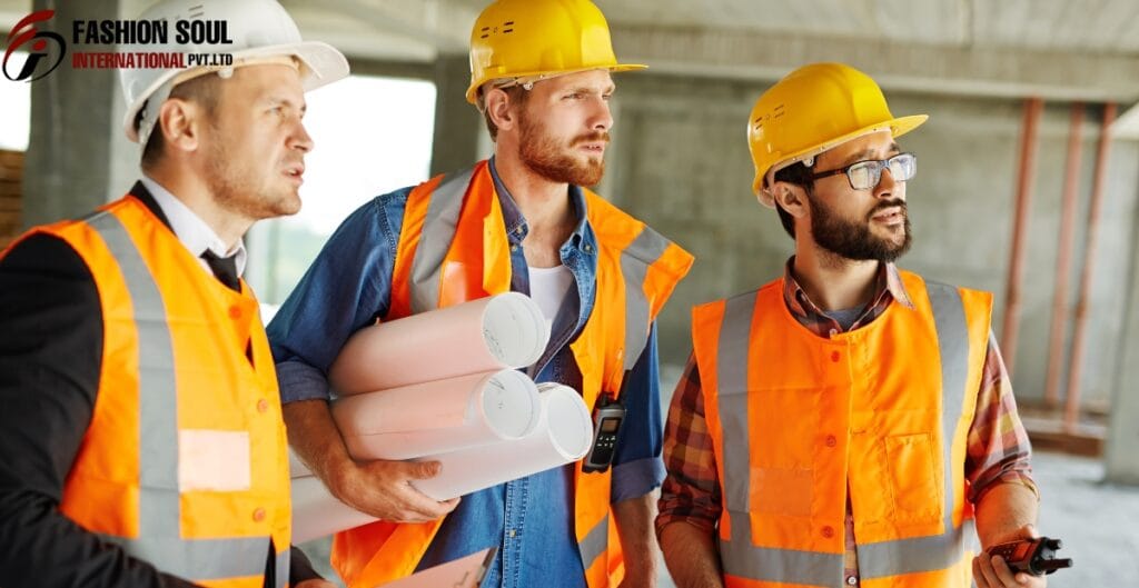 Three construction workers wearing orange safety vests and yellow helmets are standing inside a building under construction, engaged in a discussion.