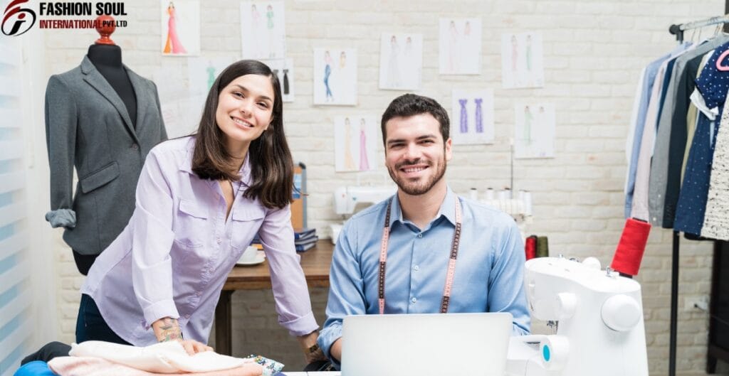 A woman and a man smiling at the camera inside a fashion design studio, with sewing equipment, fabric spools, and fashion sketches on the wall behind them.