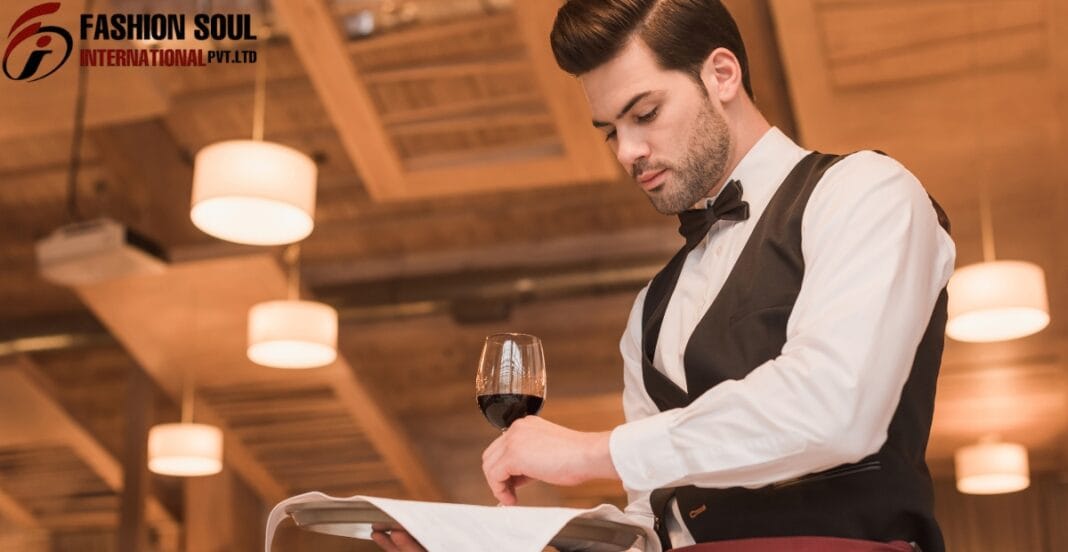 A stylish male waiter in a white shirt and black vest with a bow tie stands at a table, looking down at a menu while holding a glass of red wine. Warm dining area with soft pendant lights and wooden ceiling beams forms the cozy background; the Fashion Soul International logo is visible in the top left corner.