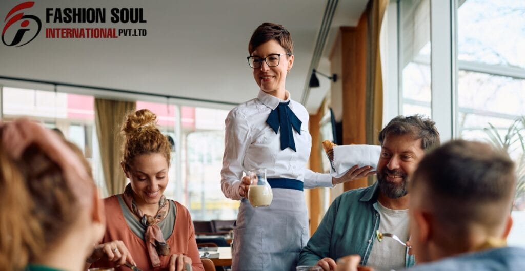 A smiling waitress in a white blouse, blue neck scarf, and gray apron serves a glass of milk to a group of diners in a bright cafe. She holds a folded napkin-wrapped bread in one hand while presenting the drink with the other.