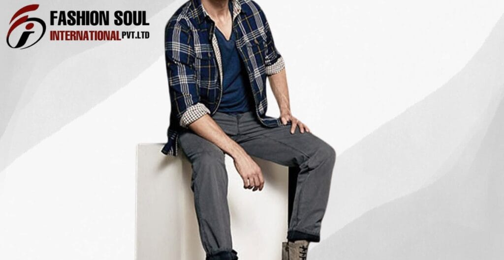 A man sitting on a white block wearing a navy and plaid shirt over a blue tee, gray pants, and tan boots. Fashion Soul International logo appears in the top left corner.