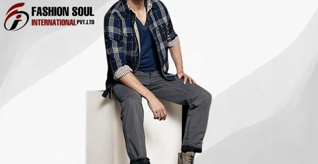 A man sitting on a white block wearing a navy and plaid shirt over a blue tee, gray pants, and tan boots. Fashion Soul International logo appears in the top left corner.