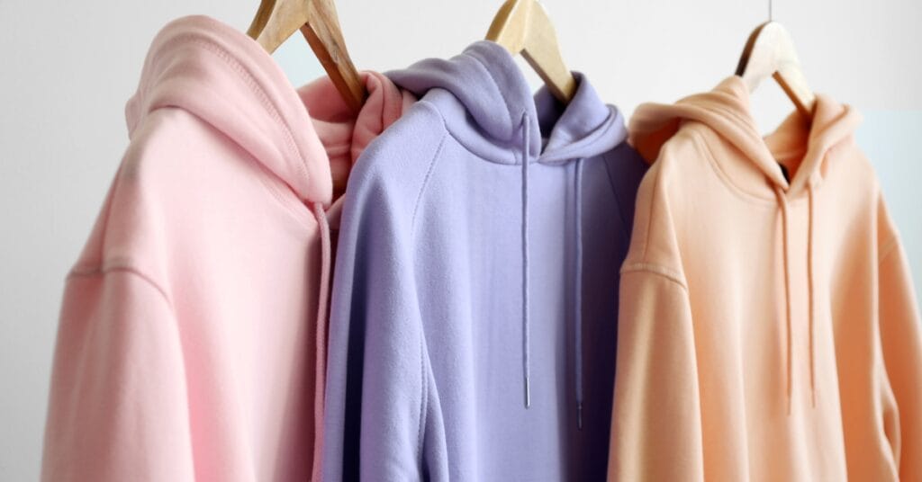 Alt text: A row of pastel-colored hoodies hanging on wooden hangers. From left to right: light pink, lavender, and peach. The hoodies feature drawstring hoods and soft, cozy fabric.