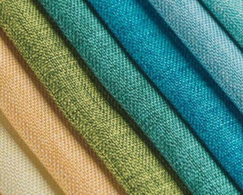  A close-up view of several folded fabrics arranged in a diagonal stack. The fabrics display a gradient of colors from warm beige and peach on the left to green and blue tones on the right, with visible textile texture and weave.