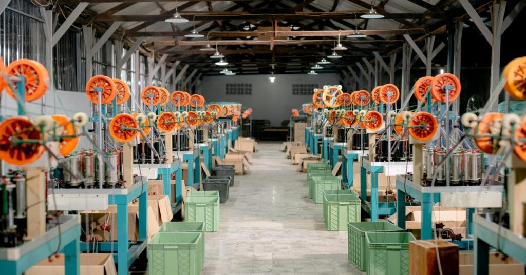 A long, well-lit workshop filled with rows of machines that have bright orange spools and teal frames. Green and brown plastic crates are placed along the aisle, and the workshop has a high ceiling with exposed beams and overhead lights.