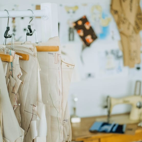 A row of sewing patterns and fabric pieces hanging on a rack in a bright workspace, with a sewing machine and sketches blurred in the background.