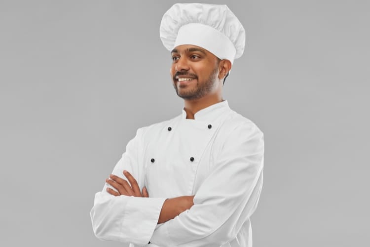 A cheerful male chef wearing a white chef’s hat and jacket, standing with arms crossed against a light gray background.