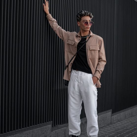 A stylish man stands against a dark, vertical slat wall. He wears a light brown oversized jacket over a black shirt, white pants, and sunglasses. One arm rests on the wall while the other hand is in his pocket, with a small black crossbody bag slung over his shoulder. The overall mood is urban and fashion-forward.