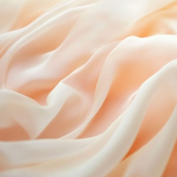 A close-up of soft, peach-toned fabric with smooth, flowing folds and a delicate, silky texture.