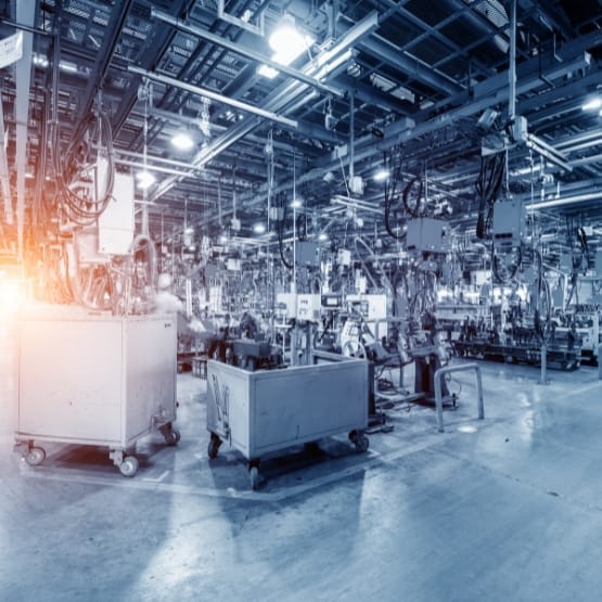 A spacious industrial factory floor filled with machinery, workstations, and overhead equipment. The scene shows a row of carts and various manufacturing stations, with bright lighting and a cool blue tone. A warm glow to the left suggests sunlight or illumination. The environment appears organized and busy, typical of a large-scale production facility.