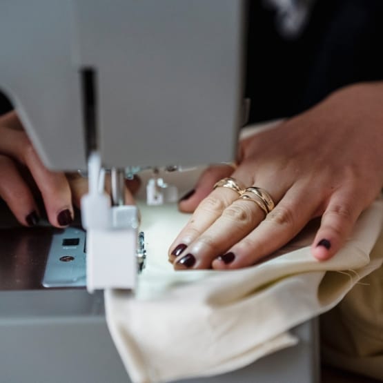 A close-up of a person sewing with a sewing machine. Their hands guide beige fabric under the machine’s presser foot; visible are dark nail polish and several rings on their fingers. The machine's needle area and feed dogs are in focus.