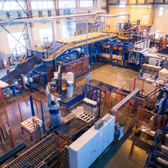 An expansive industrial manufacturing floor with automated robotic arms and conveyor belts. A multi-level blue metal framework and pathways surround the machinery, with large machines, crates, and workstations arranged in rows. The area is well-lit by natural light from windows and overhead lighting, with a clean concrete floor and organized equipment.