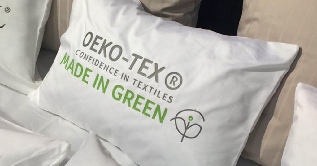 “White pillow with green text reading ‘MADE IN GREEN’ and a circular logo, brand name ‘OEKO-TEX’ on a bed.”