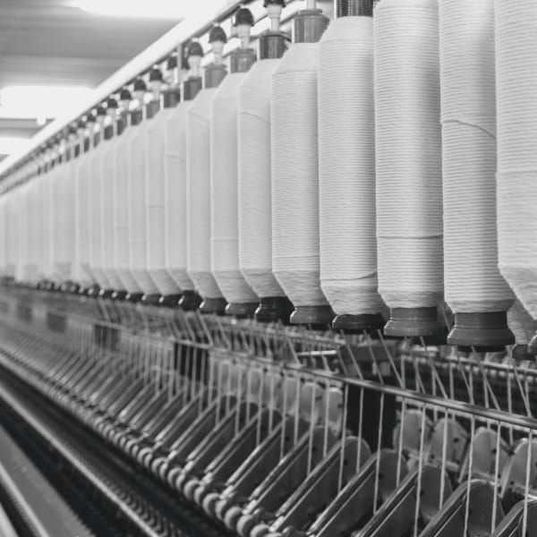A textile manufacturing machine with tall spools of white thread aligned in a row. The spools sit on a metal frame, with threads running horizontally and machinery components visible below. The scene is in grayscale, giving a high-contrast industrial appearance.