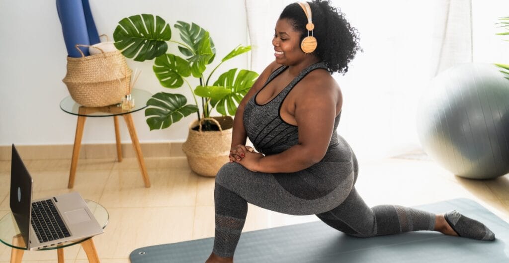 Plus size woman in a fashionable gray exercise outfit doing a low lunge yoga pose on a blue mat during a home workout, representing stylish and comfortable fitness wear.