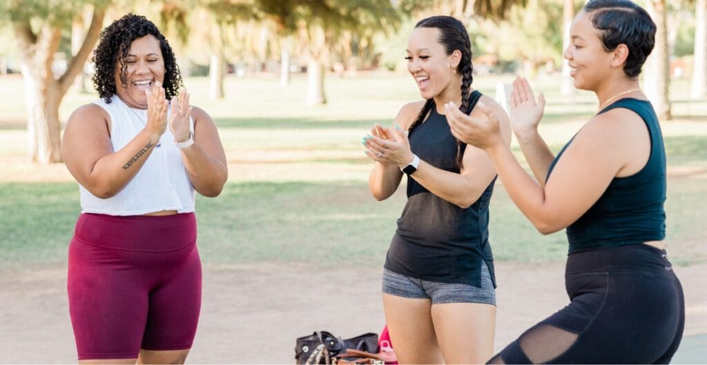Three women in plus size lycra shorts exercising outdoors, smiling, and clapping together
