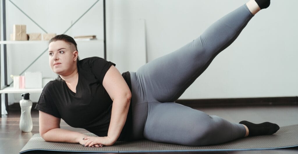 A woman doing an exercise on a yoga mat. They are lying on their side with one leg lifted high in the air and the other leg bent. They wear a black shirt and gray clothing plus size leggings, in a gym or home workout space with a water bottle nearby.