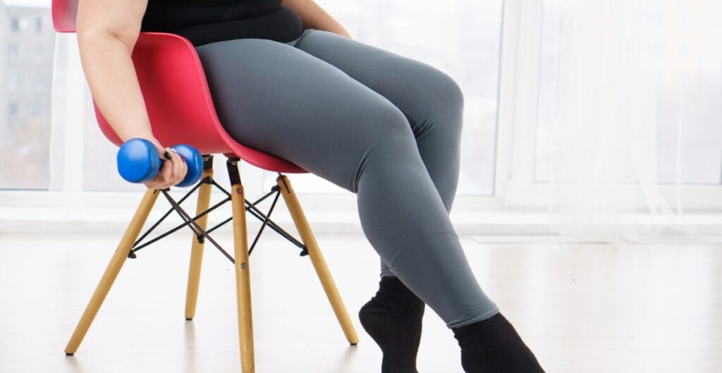 A woman sitting on a pink chair, holding a small blue dumbbell wearing plus size leggings for exercise