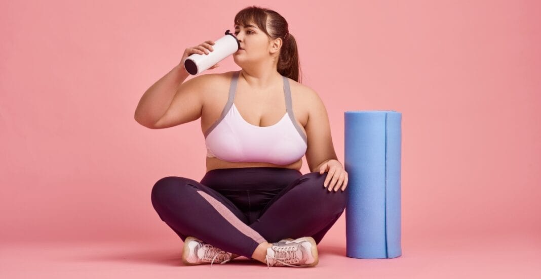 A person sits on the floor in cute plus size leggings, sipping water after a workout session.
