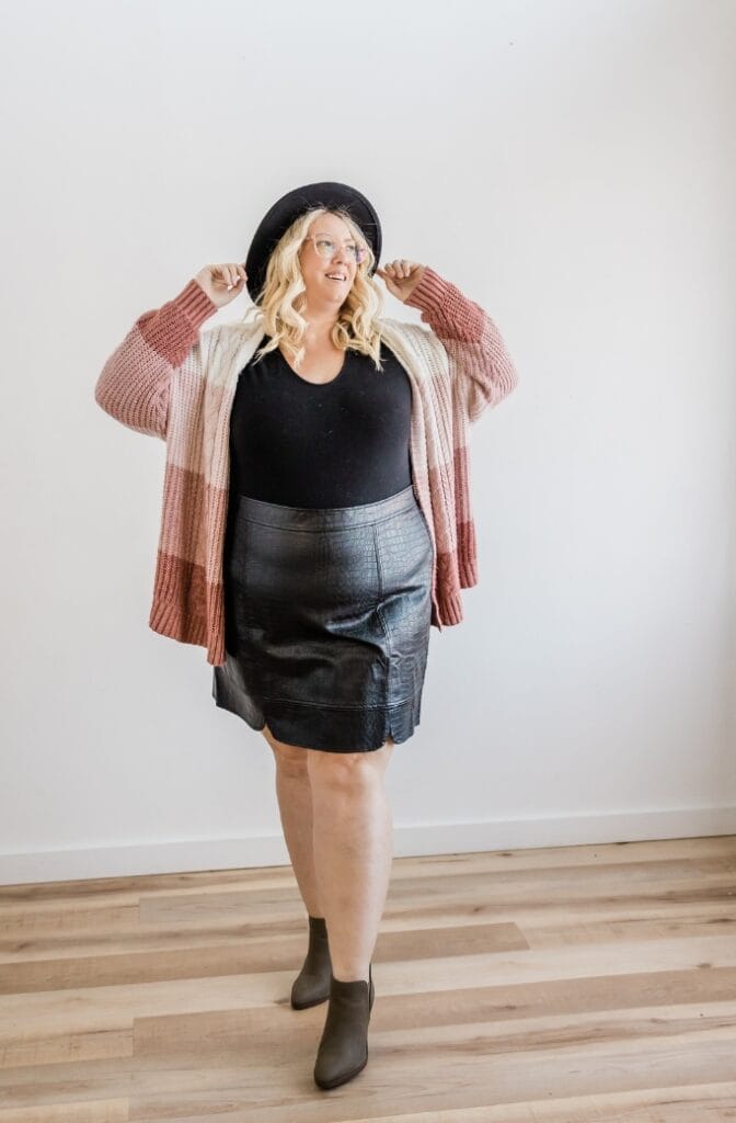 Plus size woman in a black leather skort, striped cardigan, and brown ankle boots, posing indoors with a smile