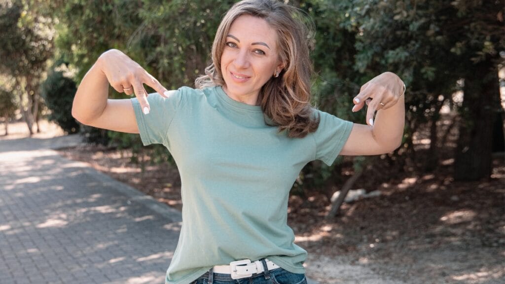 Woman smiling and pointing to her green baby doll shirt, standing outdoors in a natural setting