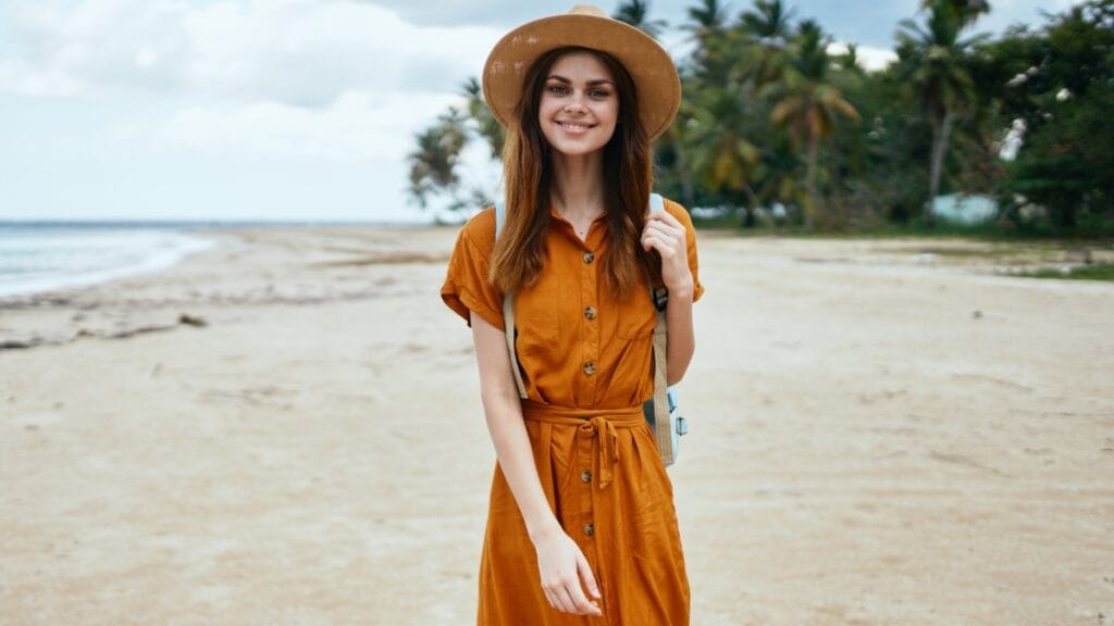 Woman wearing a mustard beach midi dress with a hat, smiling on a sunny beach.