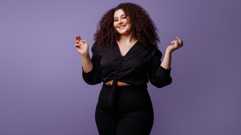 Curvy woman wearing a black crop top and black leggings, posing confidently in front of a purple background.