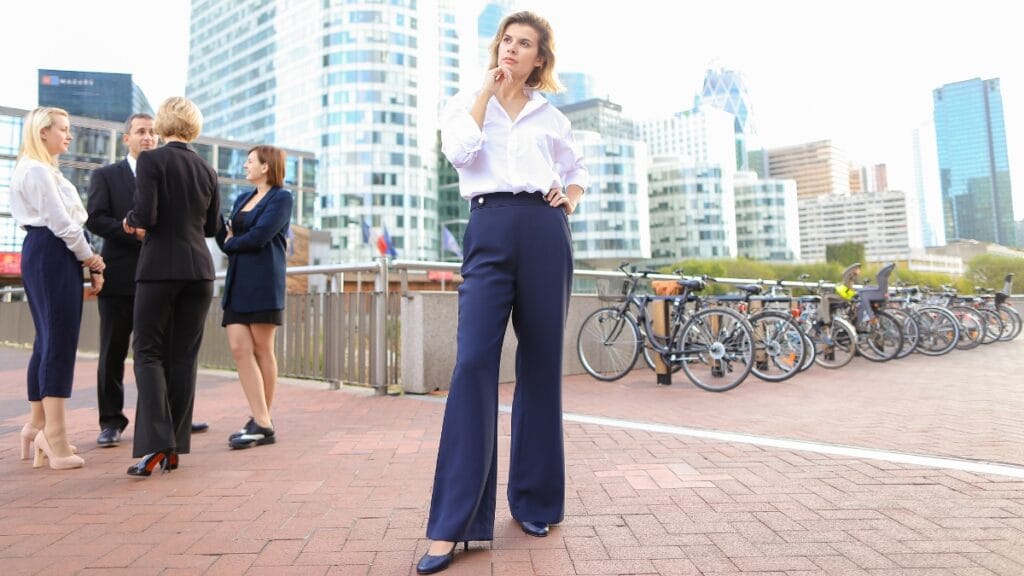 A woman confidently stands outdoors in a business setting, wearing dressy pants paired with a white blouse and high heels for a corporate look
