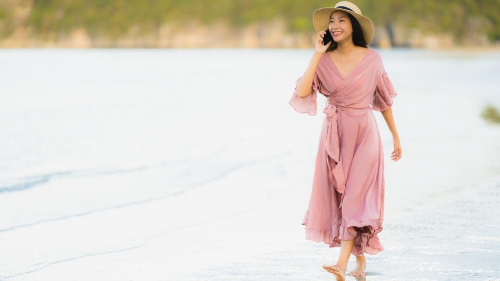 Woman in a pink flowy midi dress walking barefoot beside a calm body of water