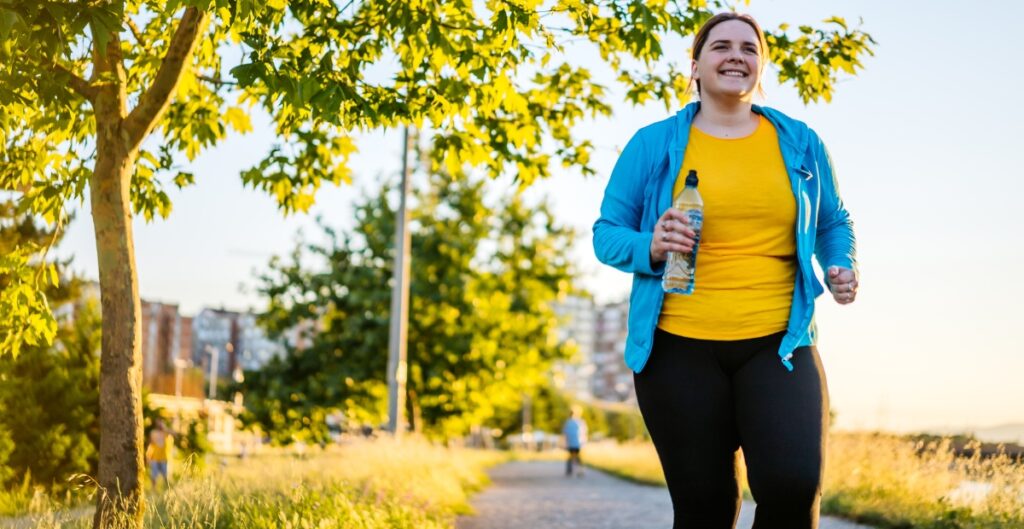 Plus size woman jogging in yellow and blue jogger suit, holding a water bottle on a sunny day.