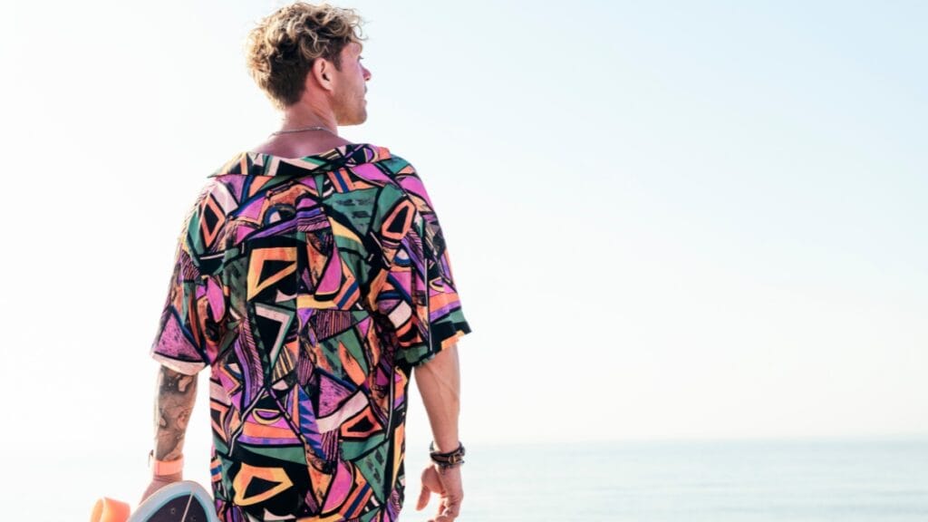 Individual wearing a vibrant 3XL graphic tee with abstract patterns, standing by the beach with a skateboard