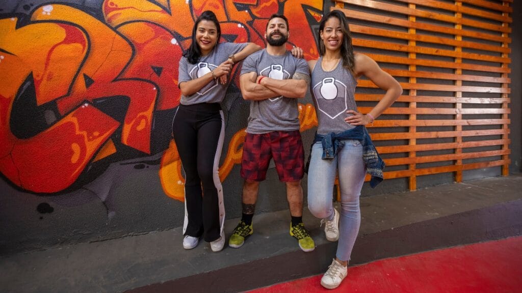 Group of three individuals wearing matching 3XL graphic tees with a fitness logo, posing in front of a graffiti wall