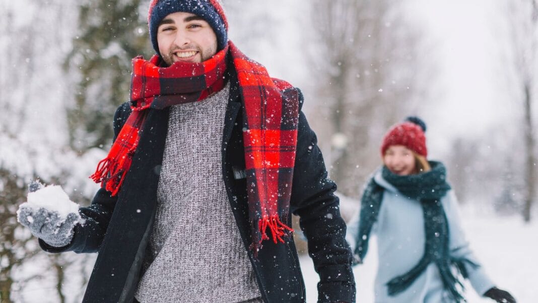 Man with a red plaid scarf and black coat holding a snowball with a woman in the background wearing a red beanie and teal scarf, walking in a snowy outdoor setting.