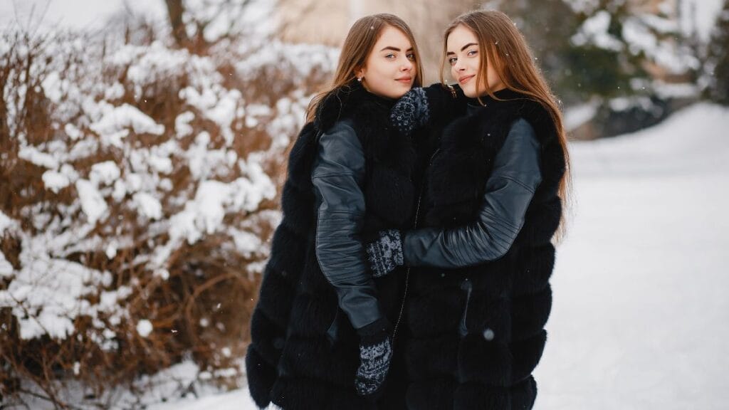 Two young women who look like twins standing close together outdoors in a snowy landscape, wearing matching black fur vests, leather sleeves, and winter gloves.