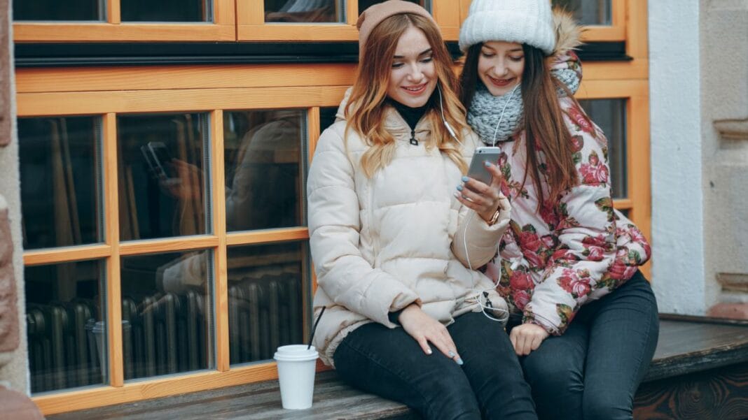 Two young women enjoying winter fashion, wearing puffer jackets and scarves while sitting on a window ledge, looking at a phone.
