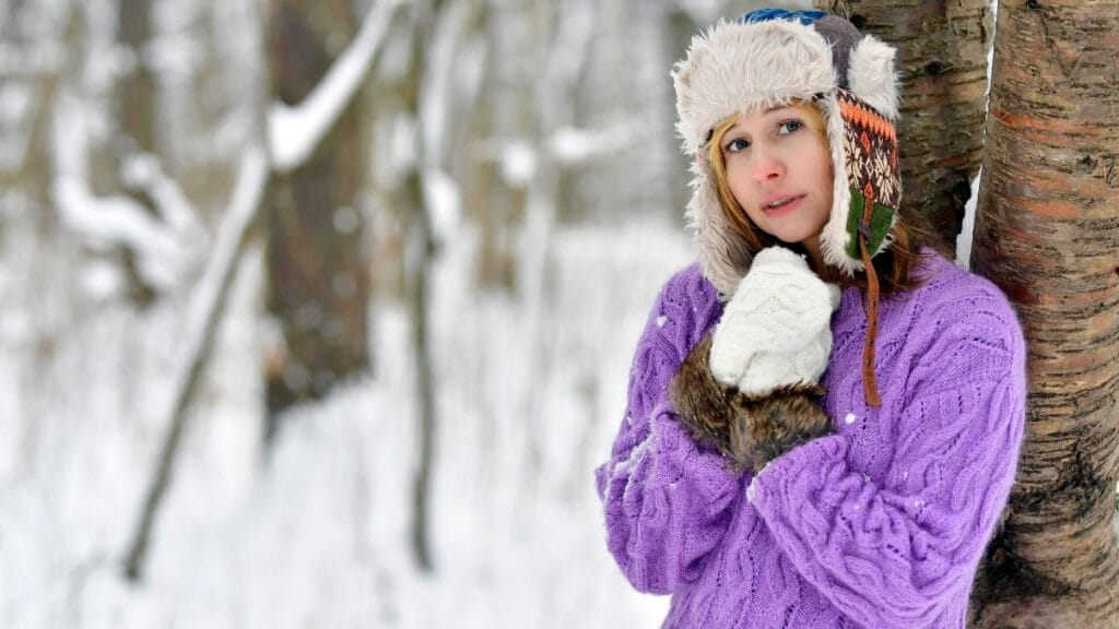 A woman wearing a purple knitted sweater, white gloves, and a warm fur-lined hat stands in a snowy forest, looking thoughtfully into the distance.