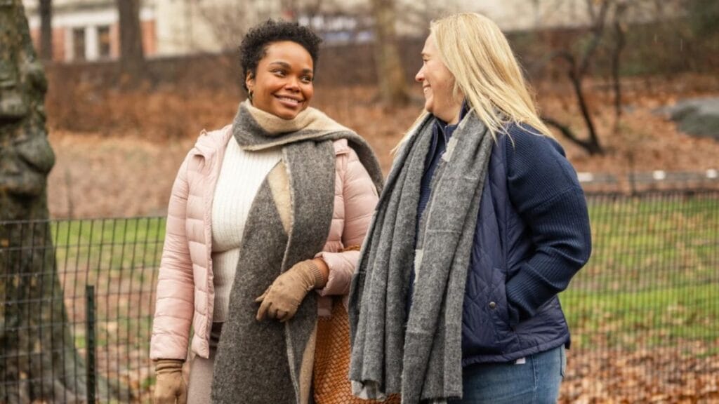 A woman standing outdoors in winter attire, wearing a long black coat, a Burberry-style scarf, a maroon sweater, and over-the-knee black boots. She is smiling and posing in front of a bushy hedge under the bright sun.