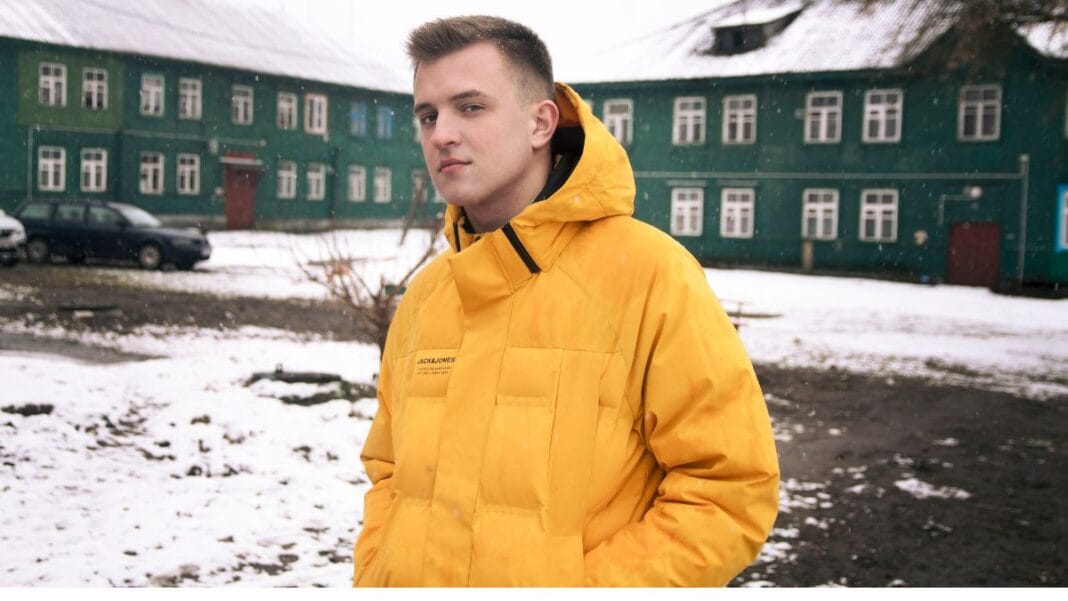 A young man stands in the snow wearing a bright yellow winter jacket, exuding confidence against a backdrop of green buildings and snowy ground.