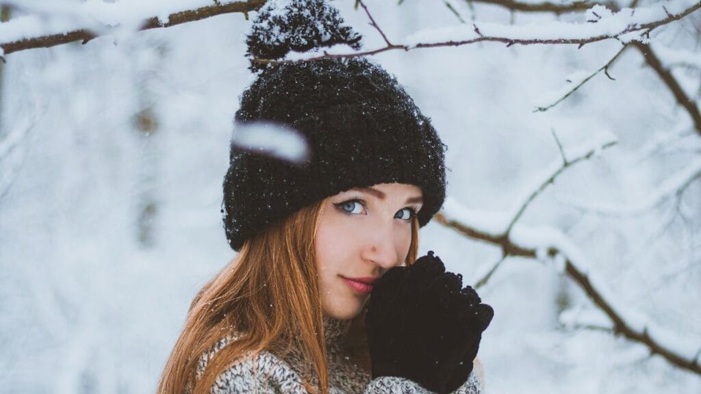 A young woman standing outdoors in a snowy forest, wearing a black knit hat and gloves, holding her hands near her face to stay warm.